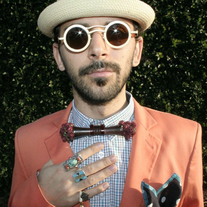 Man wearing a peach blazer, white shirt, and beige hat with sunglasses, holding a bow tie against a green hedge background.