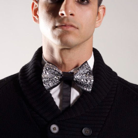 Man wearing a black coat with a white shirt and a sparkling silver rhinestone on leather bow tie on a plain background